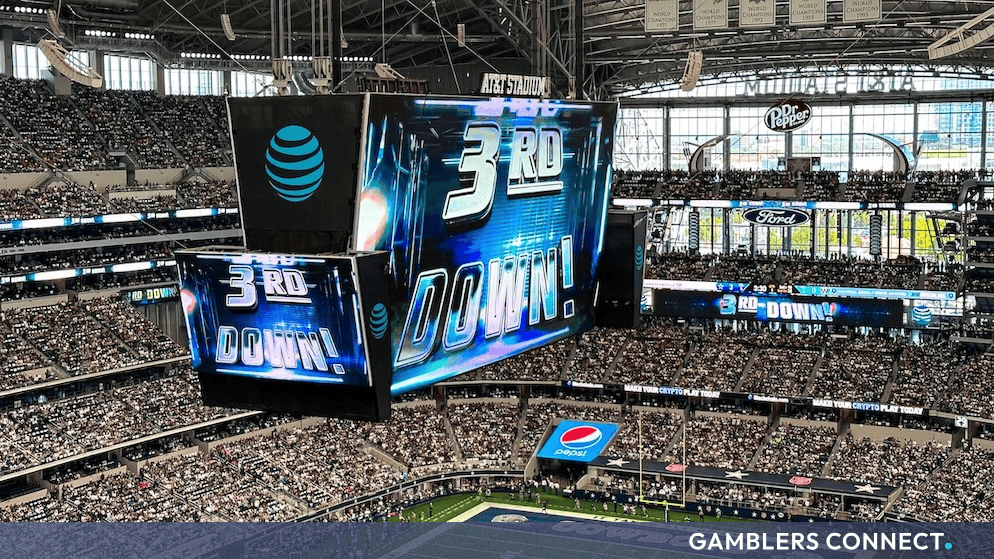 Packed American football stadium viewed from the stands, featuring a massive central replay screen displaying game action.
