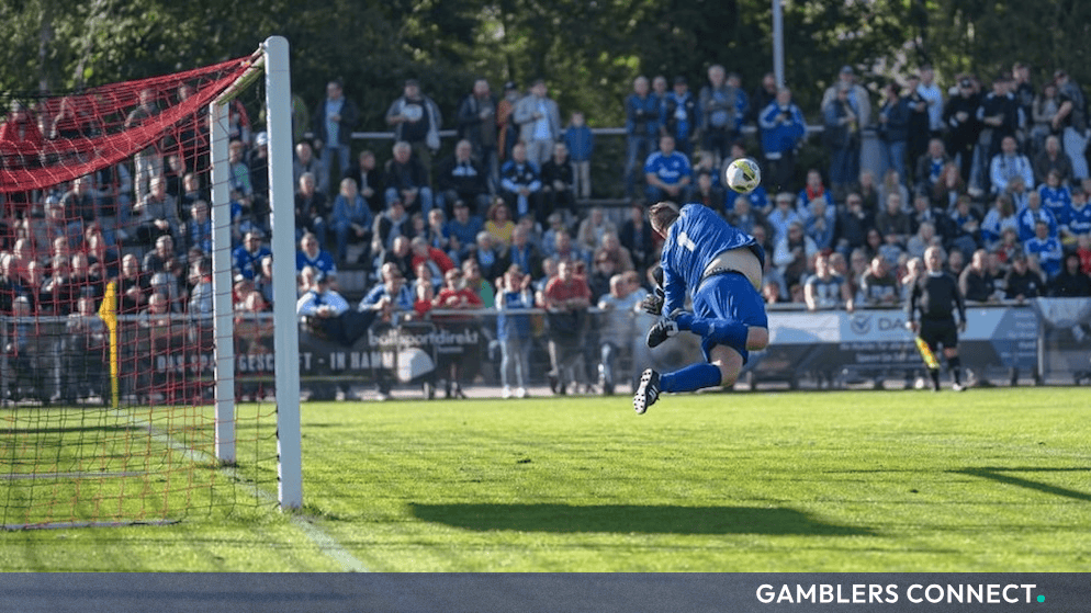A football goalkeeper diving to make a save during a lower-division match in front of spectators.