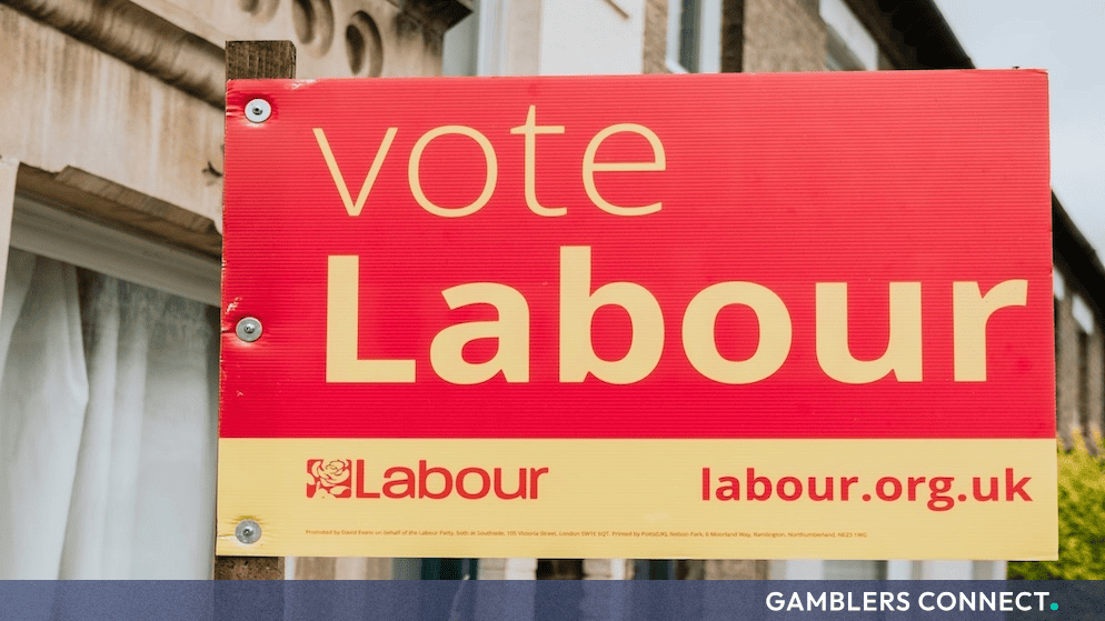 A prominent red and yellow "Vote Labour" election sign mounted on a wooden post outside a residential area in the UK.
