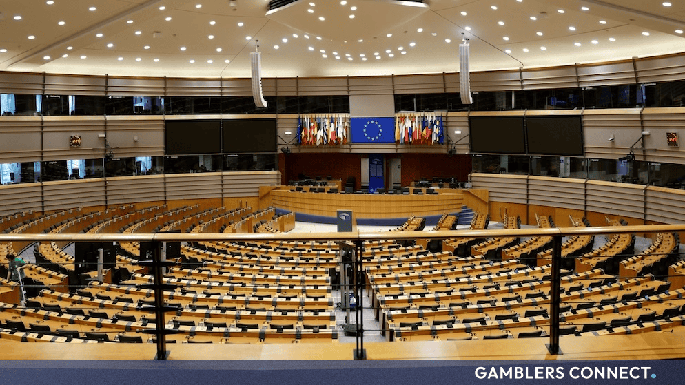 Interior view of the empty European Parliament hemicycle chamber with rows of desks and the European Union flag displayed at the front.