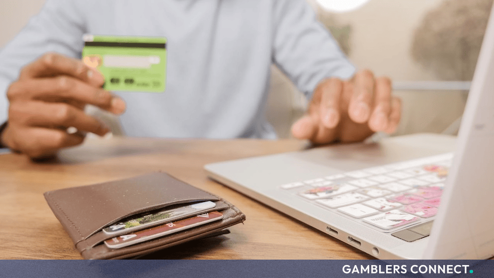 A person holding a bank card near a laptop with a wallet and cards resting on a wooden desk.