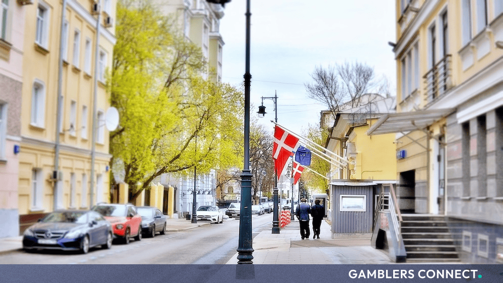 Urban street view in Denmark featuring historic European architecture, parked cars, and pedestrians walking past buildings.