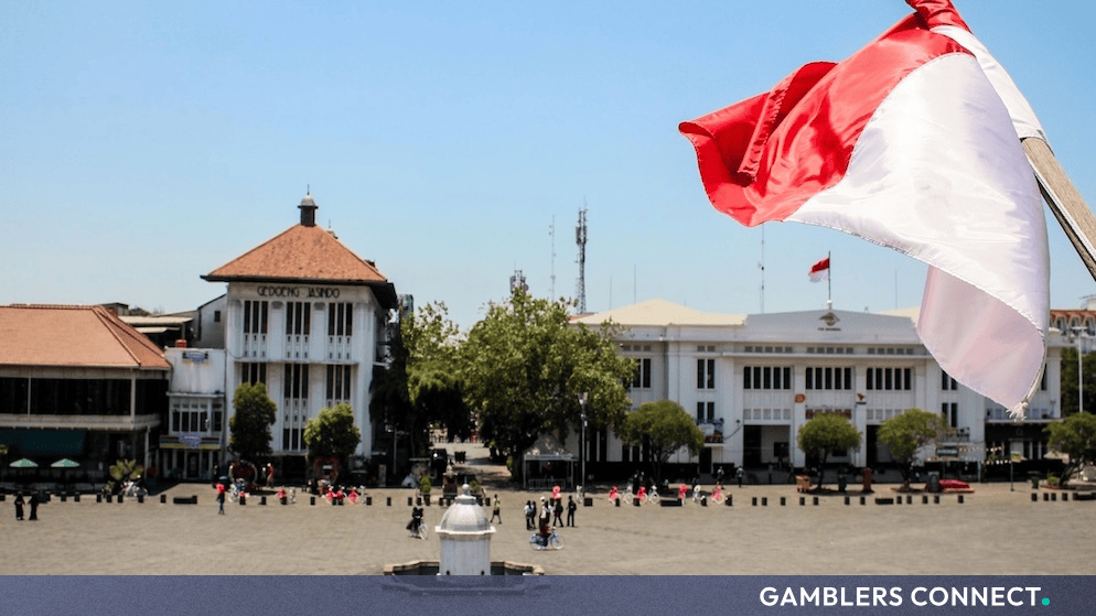 The red and white Indonesian national flag waving proudly in front of historic colonial-era architecture in a public plaza.