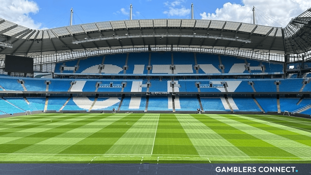 Empty blue seats inside Etihad Stadium overlooking the pitch under a bright cloudy sky.
